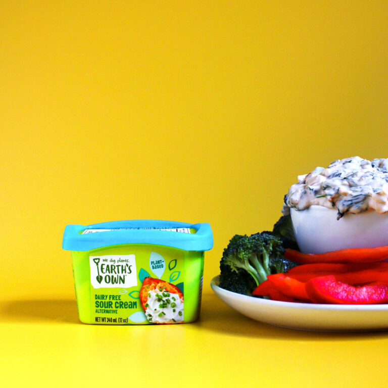 Vegan spinach dip made with earths own dairy free sour cream. Plate of veggies and sliced bread sit surrounding bowl of spinach dip.