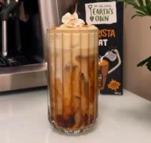 Image of pumpkin cheesecake iced latte topped with whip cream in clear glass. Carton of Oat Barista sits behind it.