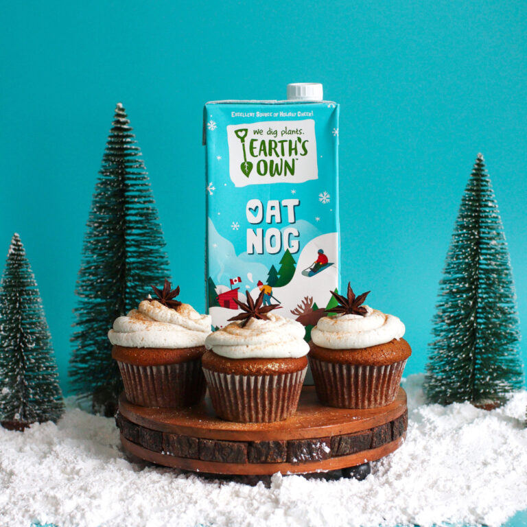 oat nog molasses cupcakes with oat nog carton surrounded by decorative christmas trees infront of blue background
