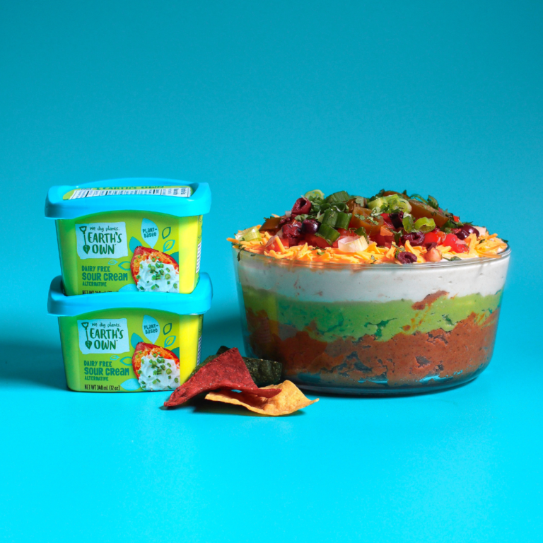 Taco Layered Dip in bowl sitting next to two tubs of Earth's Own Sour Cream